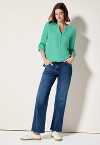 Chemisier turquoise à manches retroussées, associé à un jean bleu à jambes larges. Des baskets blanches complètent le look décontracté.