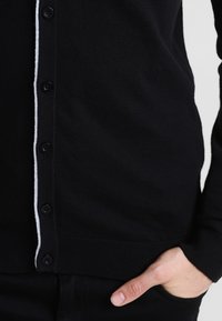 Czarny kardigan z miękkiego materiału o dopasowanym kroju. Posiada białą paskę wzdłuż listwy z guzikami oraz sześć czarnych guzików.