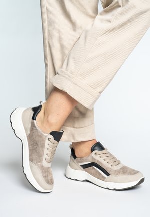 Osoba stojící v béžových manšestrových kalhotách a béžovo-černobílých teniskách se šněrováním, s jednou nohou mírně zvednutou.