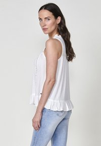Top bianco senza maniche con orlo arricciato, caratterizzato da pieghe verticali e un delicato bordo lungo la parte anteriore, abbinato a jeans azzurri chiari.