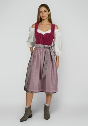 Frau trägt ein traditionelles pink-lila Dirndl mit weißer schulterfreier Bluse und grauen Stiefeletten, steht vor einem schlichten Hintergrund.