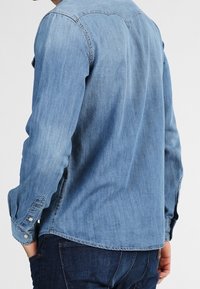 Chemise en denim bleu clair avec des manches longues, des poignets à boutons et un ourlet arrondi. Dotée d'un col classique et d'un léger dégradé sur le tissu.