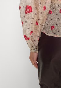 Durchsichtige beiger Bluse mit roten Blumenmustern, Punkten und einer strukturierten Oberflächenbeschaffenheit. Die aufgekrempelten Ärmel verfügen über einen silbernen Hardware-Detail.