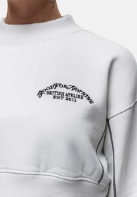 Biała bluza z okrągłym dekoltem, z czarnym haftowanym napisem "Good For Nothing British Atelier Established 2011" na klatce piersiowej.