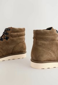 Botas de tobillo de ante marrón con cordones en la parte delantera, puntera redonda y suela de goma blanca. Presenta detalles de costura y un cierre con cremallera en la parte posterior.