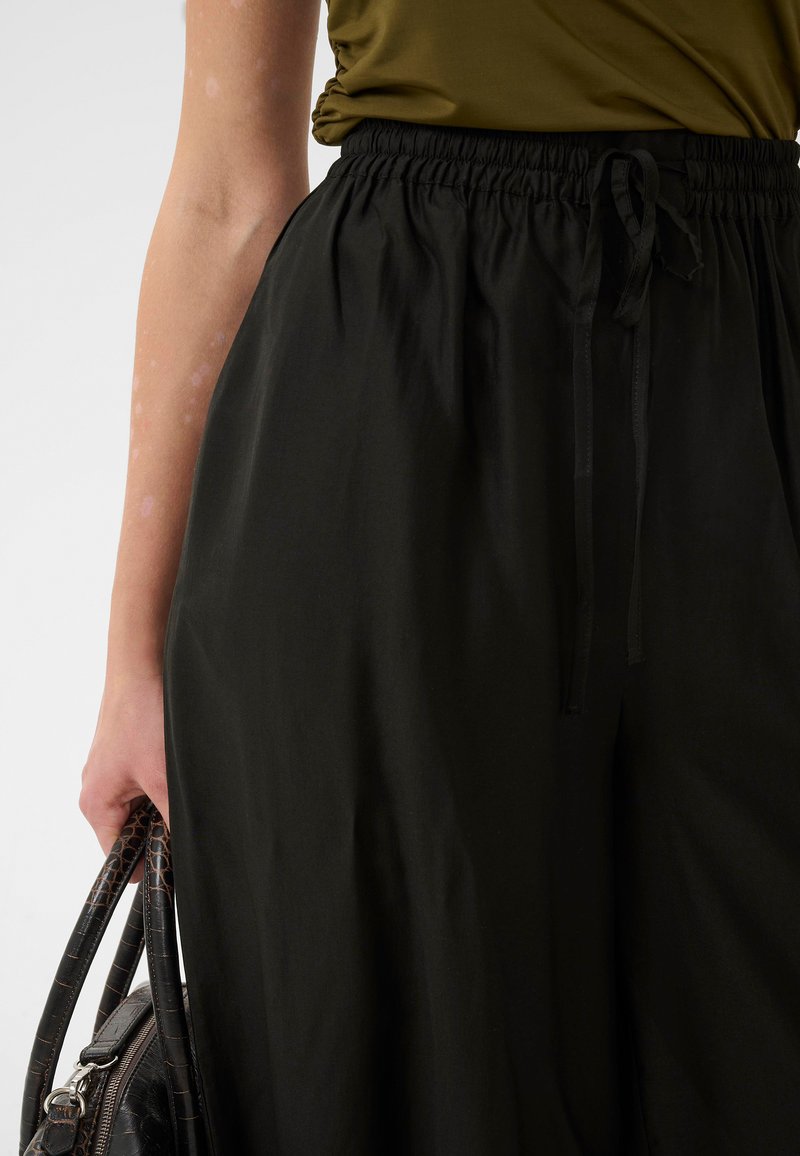 Personne portant une chemise vert olive et un pantalon noir à taille élastique, tenant un sac à main noir texturé à ses côtés.