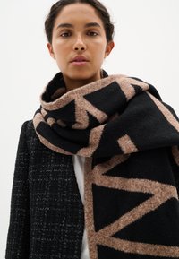 Svart och brun mönstrad scarf i mjuk, strukturerad ull, med geometriska mönster, hängande runt halsen och bärs med en svart jacka.