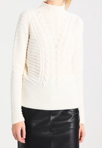 Weißer Strickpullover mit hohem Kragen, der über ein Kabelstrickdetail und gerippten Textur am Saum und an den Ärmeln verfügt. Kombiniert mit einem schwarzen Lederrock.