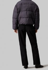 Osoba ubrana w czarną pikowaną kurtkę, czarne proste spodnie dżinsowe i czarne botki na obcasie stoi na tle jednolitego szarego tła.