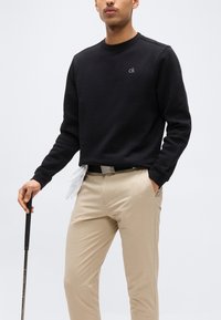 Homme portant un sweat-shirt noir Calvin Klein et un pantalon beige, tenant un club de golf avec un gant blanc glissé dans la ceinture.
