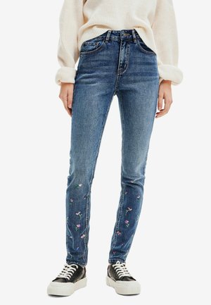 Kvinde iført blå skinny jeans med små broderede blomster nær anklerne, en cremefarvet trøje og sorte sneakers med hvide såler.