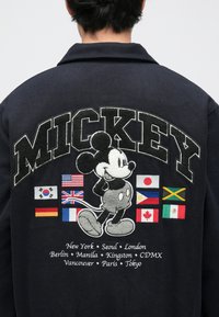 Casaco preto com design bordado do Mickey Mouse, bandeiras de países e texto com nomes de cidades, com gola e tecido texturizado.