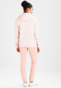 Jachetă de vânt roz deschis, cu glugă, manșete elastice și o croială lejeră, asortată cu pantaloni jogger roz și adidași albi.