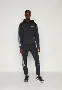 Hoodie noir avec des rayures turquoises sur les manches, arborant un logo. Jogging noir avec des rayures blanches sur les côtés. Chaussures de sport noires.