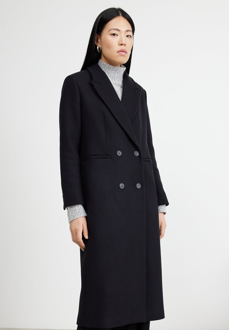 Cappotto nero doppiopetto con silhouette strutturata, realizzato in misto lana, con grandi bottoni e tasche laterali. Sotto, un dolcevita grigio strutturato.
