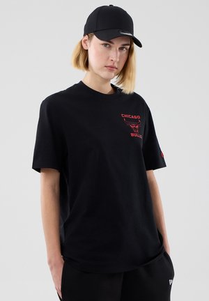 Młoda osoba z blond włosami, ubrana w czarną czapkę i czarną koszulkę Chicago Bulls, stoi na tle jednolitego jasnego tła.