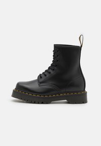 Stivale alla caviglia in pelle nera con punta rotonda, cuciture gialle, chiusura con lacci frontale e suola in gomma strutturata. Presenta una linguetta sul retro.