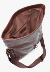 Brown leather tote bag with a soft texture, large interior, and a zippered pocket. Features a wide fabric strap with metal accents.