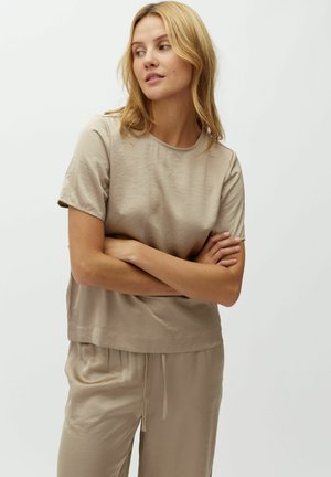 Femme aux cheveux blonds portant un haut beige à manches courtes et un pantalon assorti, les bras croisés, regardant sur le côté devant un fond uni.