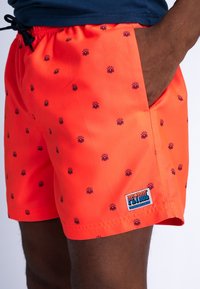 Shorts de baie portocalii strălucitoare cu modele mici de soare și valuri în nuanță de navy, având un brâu elastic și o buzunar lateral. Eticheta brandului este vizibilă.