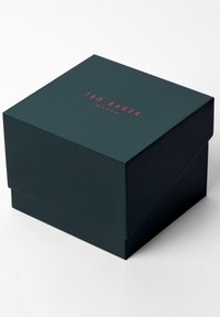 Caja cuadrada de color verde oscuro con una tapa, que presenta el texto rojo "TED BAKER LONDON" en la parte superior. Acabado suave y mate con esquinas afiladas.