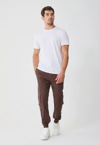 Biała koszulka z okrągłym dekoltem w stylu crew-neck w połączeniu z brązowymi dresowymi spodniami cargo z bocznymi kieszeniami, elastycznymi mankietami oraz białymi sneakersami na neutralnym tle.