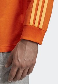 Sudadera de algodón naranja con tres rayas verticales amarillas en la manga; puño acanalado texturizado; cinturilla gris de los pantalones adyacentes visible.