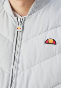 Ljusgrå bomberjacka med kviltad design, ribbat krage och dragkedja. Har en broderad logotyp med röda och gula accenter.