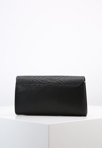 Černá obdélníková psaníčko s texturovaným klopovým víčkem v hadím vzoru, umístěné na bílé geometrické podstavě.