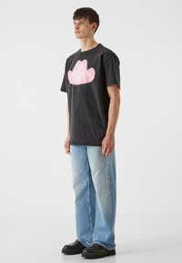 Melna pārmērīga T-krekls ar rozā mākoņa formas grafiku uz priekša, apvienots ar gaiši ziliem taisnaviem džinsiem un melniem masīviem apaviem.