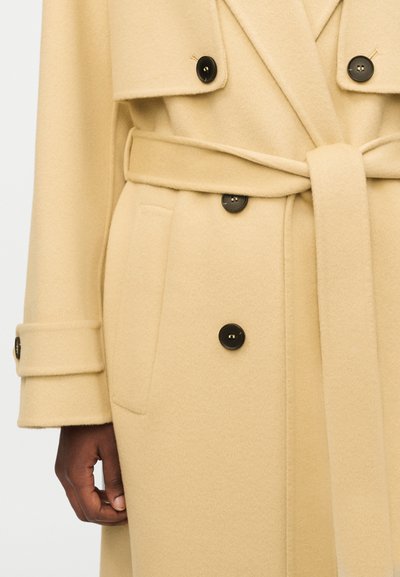 Μπεζ παλτό από μάλλινο ύφασμα με ζώνη, μαύρα κουμπιά, λουράκι στο μανίκι και πλαϊνή τσέπη· αριστερό χέρι μοντέλου ορατό στο κάτω μέρος του παλτού.
