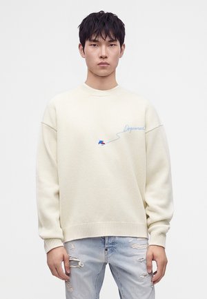 Jeune homme aux cheveux noirs porte un pull crème avec broderie bleue et un jean clair déchiré, debout sur un fond blanc uni.