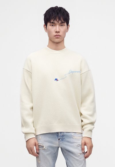 Jeune homme aux cheveux noirs porte un pull crème avec broderie bleue et un jean clair déchiré, debout sur un fond blanc uni.