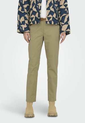 Beige quilted jacket with geometric navy patterns, paired with olive green tapered pants and beige ankle boots. Smooth fabric and fitted design.