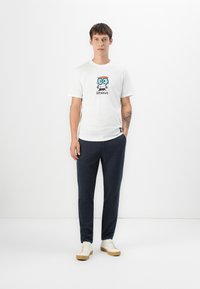 Ung mand står, iført hvide sneakers, mørke bukser og en hvid T-shirt med en tegneseriefigur og teksten "Genius" på.