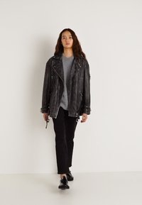 Melna ādas biker stila jaka ar sudraba rāvējslēdzējiem, uzvilkta virs pelēkas T-krekla, kombinēta ar melnām biksēm un melniem apaviem, uz minimālistiska fona.