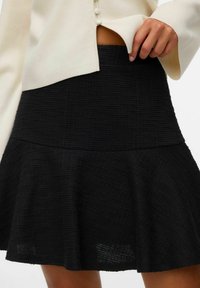 Jupe noire texturée avec un ourlet évasé et une taille ajustée. Le matériau semble tissé avec un motif subtil. Un haut blanc boutonné est partiellement visible.