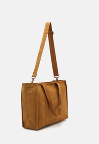 Bolsa de lona marrón con dos asas y una correa de hombro desmontable. Forma rectangular, fondo plano y detalles de costura visibles.