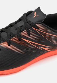Zapatilla atlética negra con superficie texturizada, acentos en naranja y un diseño distintivo con líneas diagonales; cordones negros y suela de goma.