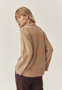 Maglione beige lavorato a maglia con colletto alto a costine e maniche lunghe, caratterizzato da una vestibilità comoda e orlo a costine, abbinato a pantaloni marrone scuro.