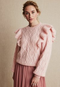Jasnoróżowy sweter z dzianiny z wzorem w warkocze, falbankowymi rękawami i okrągłym dekoltem. Miękka tekstura i luźny krój.