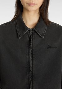 Zwarte denim jas met een puntige kraag, voorzipper en subtiele geborduurde logo op de linkerborst. Kenmerkt zich door een gestructureerde, licht versleten stof.