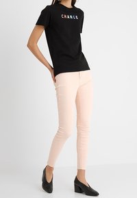 Camiseta negra con la palabra "CAMBIO" en letras coloridas, combinada con jeans ajustados color rosa claro y zapatos de tacón negros. Diseño sencillo, ajuste casual.
