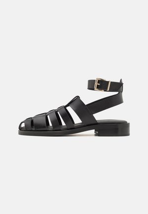 Black leather sandals featuring a strappy design, open toe, and an ankle strap with a gold buckle. Flat sole with minimal texture.