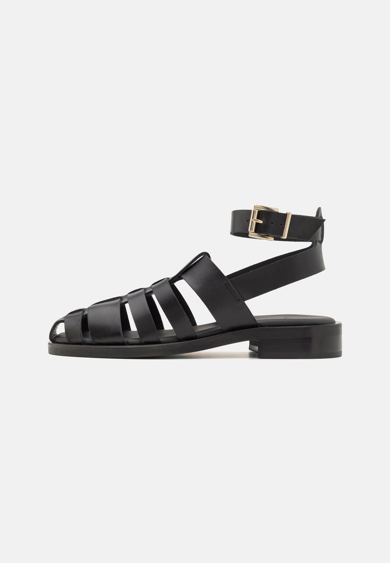 Sandali in pelle nera con un design a strisce, punta aperta e una fascia alla caviglia con una fibbia dorata. Suola piatta con texture minimale.