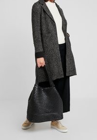 Sac fourre-tout noir tissé avec une finition texturée, présentant une forme souple et des poignées allongées, associé à un long manteau gris et des baskets beige.