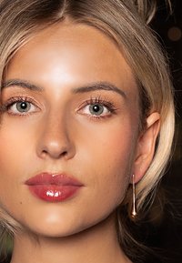 Gold drop earrings hang from the model's ear, complementing her lightly textured, pale complexion and glossy, nude lipstick.