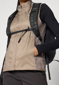 Beżowa, lekka kamizelka z wysokim kołnierzem i pełnym zamkiem, z niebieskimi akcentami, w połączeniu z czarną bluzą z długim rękawem i regulowanymi czarnymi paskami plecaka.