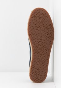 Semelle d’une chaussure avec texte en relief répété "TOMMY HILFIGER" en matériau caoutchouc marron sur fond blanc.