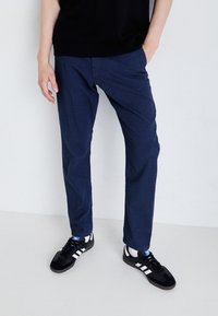 Osoba v modrých kalhotách, s černými teniskami Adidas se bílými pruhy a černým topem, stojící s jednou rukou v kapse na bílém pozadí.